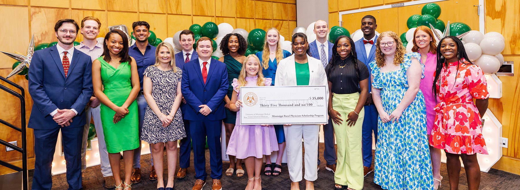 Group standing indoors holding an oversized scholarship check, with green and white balloon decorations and illuminated letters behind them.